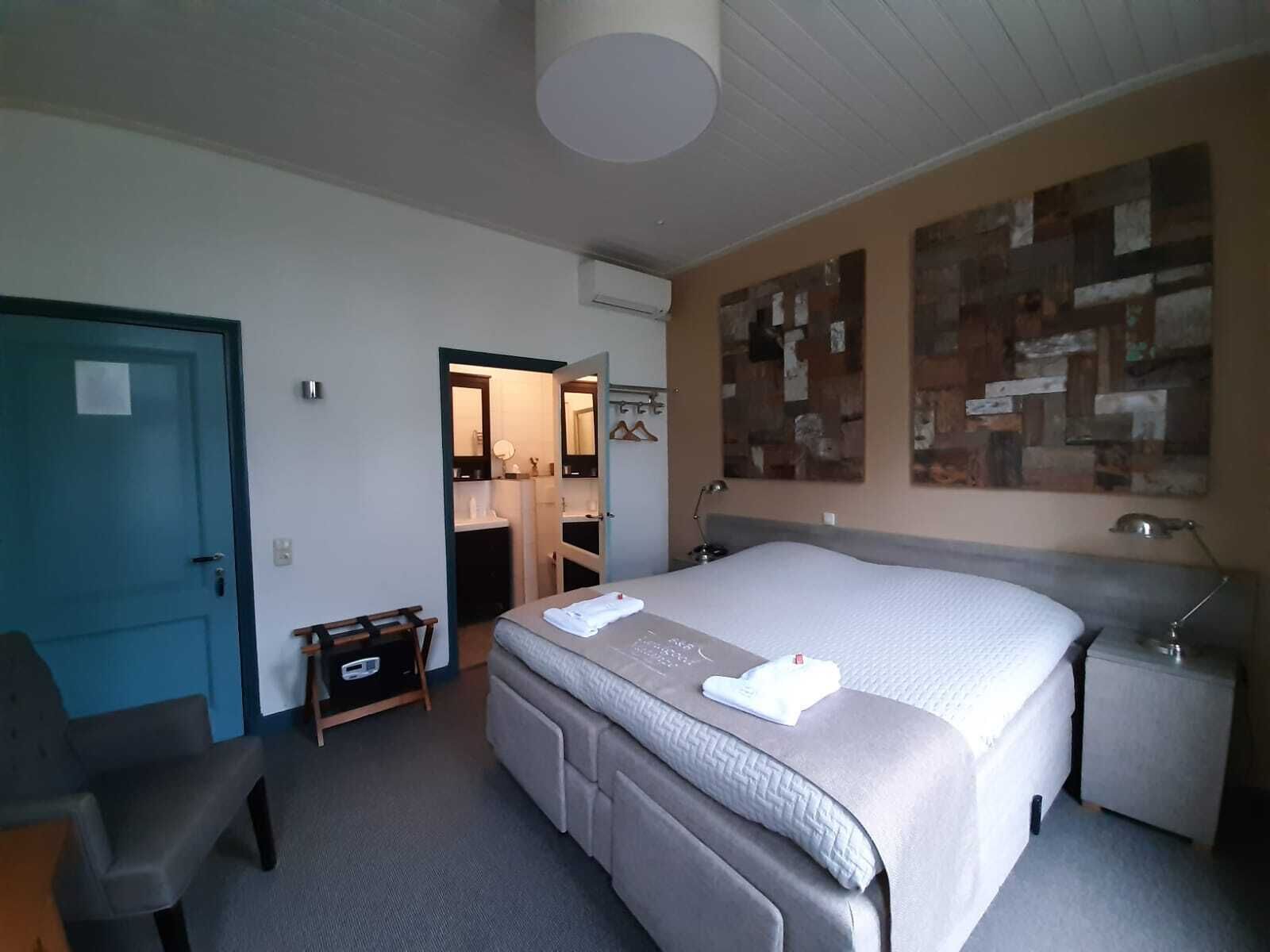 Een slaapkamer met een bed, nachtkastje, stoel, kast en een zichtbare badkamer door een geopende deur.