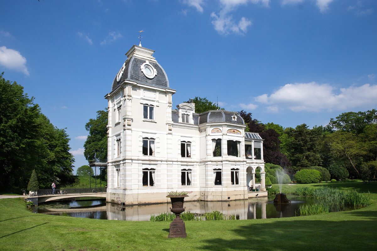 Een groot, historisch gebouw met een opvallende klokkentoren, omgeven door een verzorgde tuin en een kleine vijver.