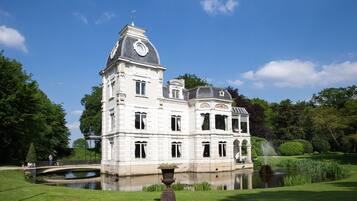 Een groot, historisch gebouw met een opvallende klokkentoren, omgeven door een verzorgde tuin en een kleine vijver.