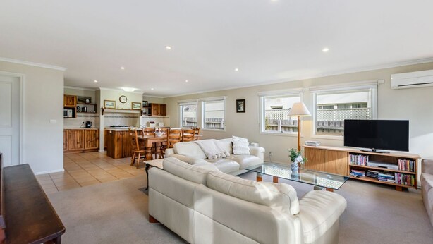 A spacious living area with a sofa, a glass coffee table, and a TV stand with books. There is a kitchen in the background with wooden cabinets and a dining area with chairs. - Centrally located & lovingly maintained family home (PORT FAIRY)