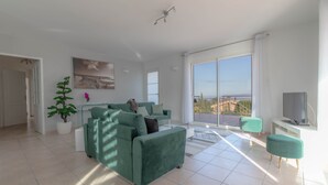 Un salon spacieux avec un canapé vert, une table basse et une plante en pot. Une grande fenêtre offre une vue sur la ville.