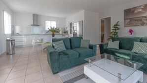 Un salon moderne avec un canapé vert, une table basse en verre et une cuisine en arrière-plan.