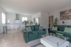 Un salon moderne avec un canapé vert, une table basse en verre et une cuisine en arrière-plan.