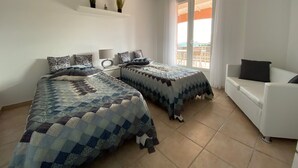 Une chambre avec deux lits, un canapé blanc, une petite table avec une lampe et une fenêtre avec des rideaux.