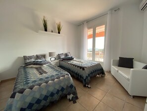 Une chambre avec deux lits, un canapé blanc, une petite table avec une lampe et une fenêtre avec des rideaux.