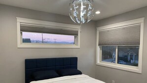 Un dormitorio con una cama, cabecera azul oscuro, dos ventanas con persianas y una lámpara de araña moderna.