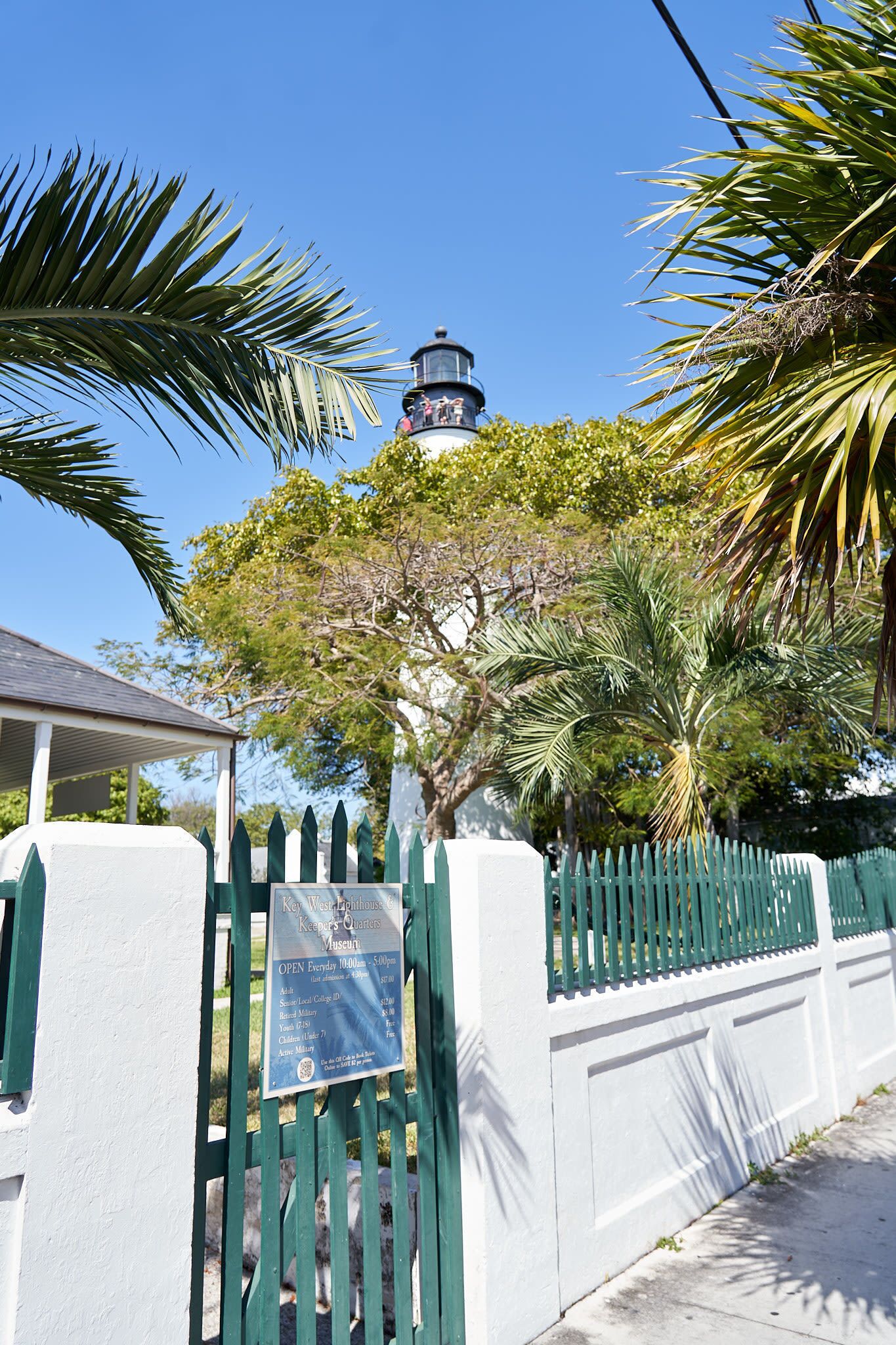 A coastal view with a lighthouse, palm trees, and a white fence with a sign.