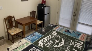 Una habitación pequeña con una mesa y sillas de madera, un refrigerador negro, un colcha en el suelo y una puerta con persianas.