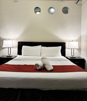Een net opgemaakt bed met witte lakens en een rode deken, omringd door twee nachtkastjes met lampen, en drie spiegels aan de muur erboven.