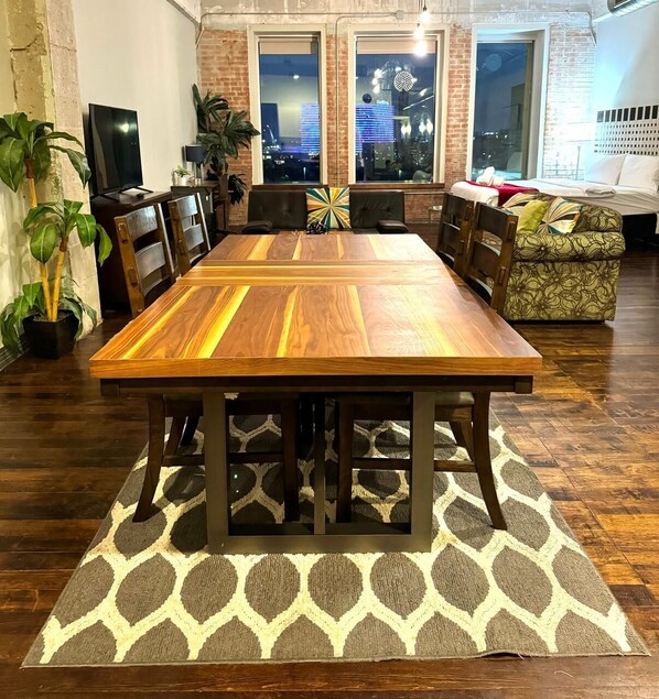 Een eetruimte met een grote houten tafel, stoelen, een kamerbreed tapijt en uitzicht op de stad door grote ramen.