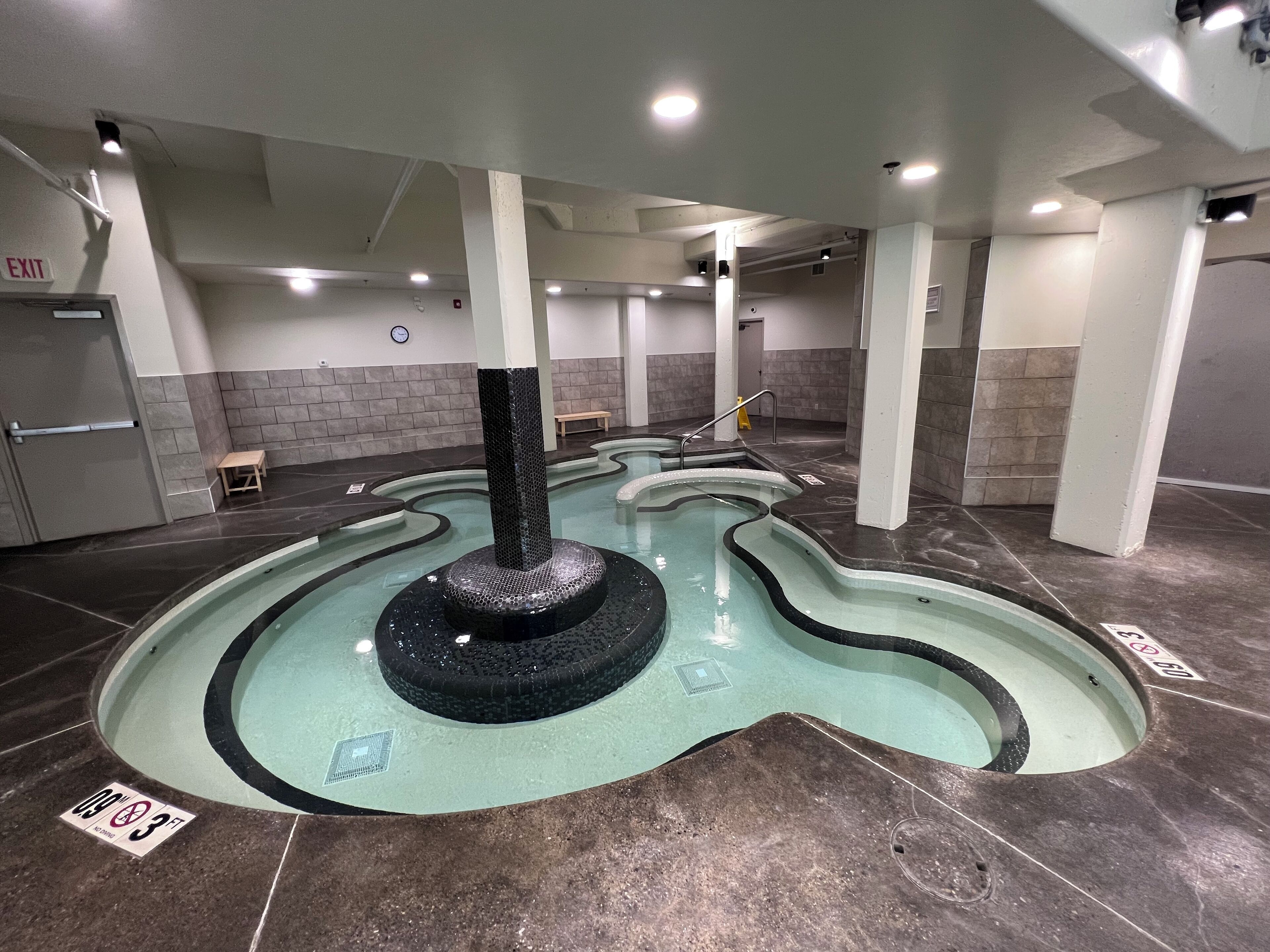 A swimming pool with a central fountain, tiled walls, and an exit sign.