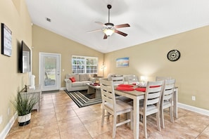 A spacious living area with a dining table, chairs, a sofa, a ceiling fan, and a large window.