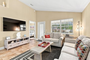A living room with a flat-screen TV, a coffee table, a sofa, and a window with a view of greenery.