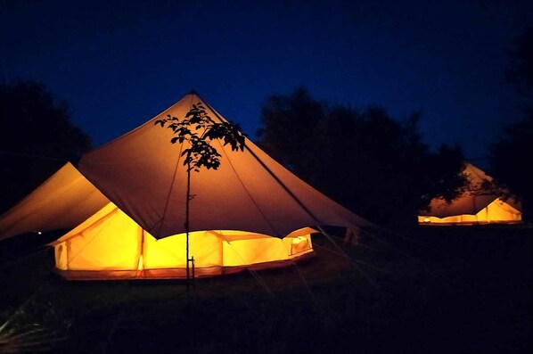 A large, illuminated tent with a conical roof structure, surrounded by trees.
