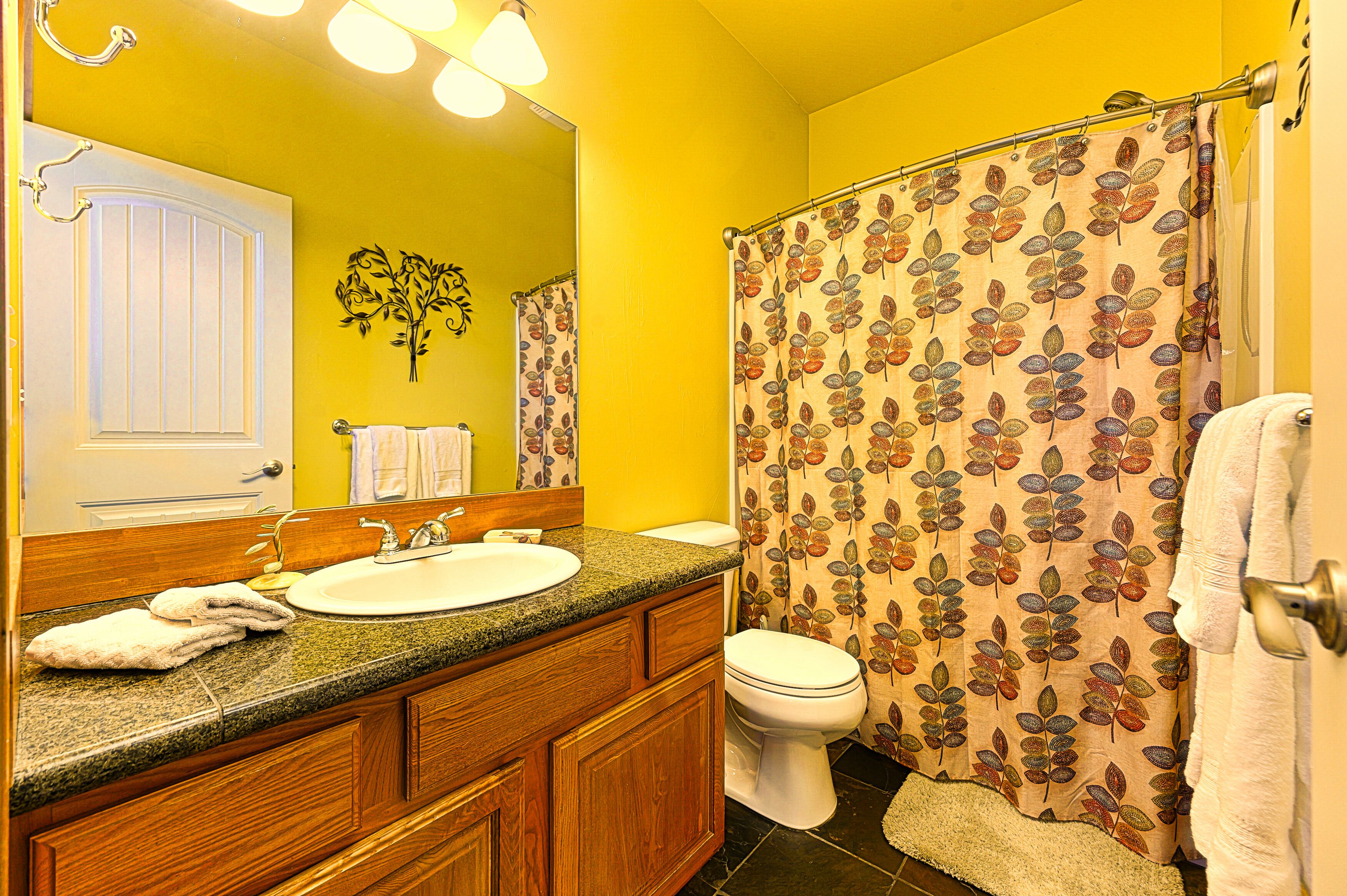 Un baño con pared amarilla, tocador de madera, lavamanos, inodoro y cortina de ducha con estampado de hojas.