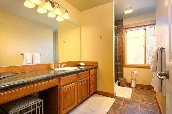 A bathroom with a wooden vanity, a large mirror, a shower with a glass door, and a window providing natural light. - Smack Dab in the Middle of Bozeman! (Bozeman)