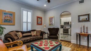 Ein geräumiges Wohnzimmer mit Sofa, Sessel und Couchtisch, im Hintergrund eine Küche und ein Deckenventilator.