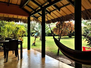 A covered patio with a hammock, a table with chairs, and a view of a well-maintained garden with various plants and a person walking in the background. - La Casita! (Las Galeras)