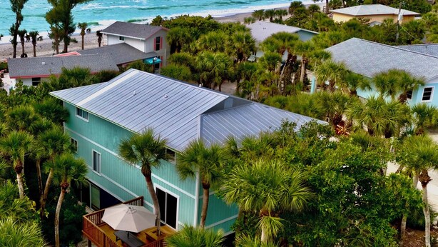 Une maison turquoise à deux étages, dotée d’un patio couvert, entourée de palmiers et offrant une vue sur la plage.