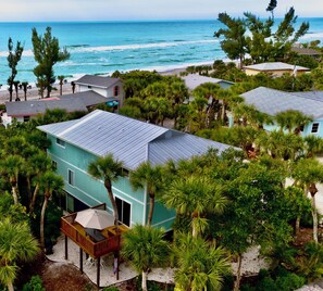 Een turquoise huis met twee verdiepingen, met een overdekte patio, omringd door palmbomen en uitzicht op het strand.