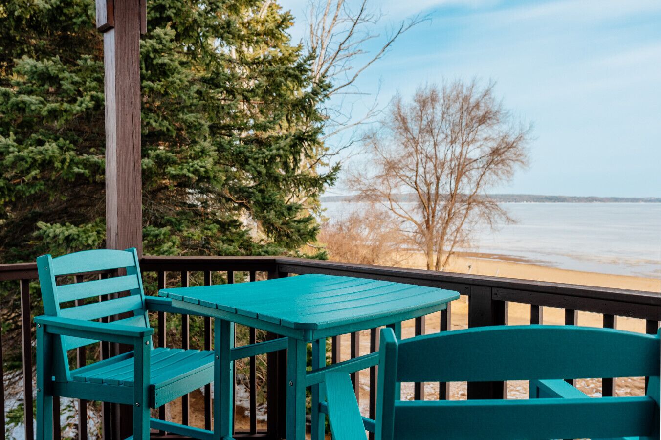 A wooden deck with a table and chairs, overlooking a beach and trees.