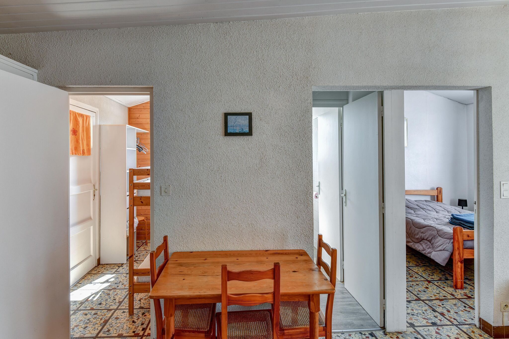 Een kamer met een houten eettafel en stoelen, een klein, ingelijst beeldje aan de muur, en een bed met een houten hoofdbord in de aangrenzende ruimte.
