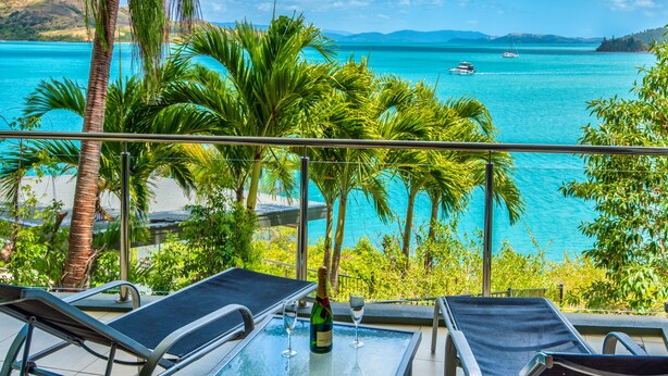 A balcony with two lounge chairs, a table with a bottle and glasses, and a view of a turquoise sea with boats. - Edge 10 - Magnificent Views! (Hamilton Island)
