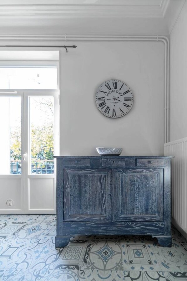 Una habitación con un reloj antiguo en la pared, un armario de madera azul, baldosas de suelo con diseño y un ventanal con vistas a zonas verdes.