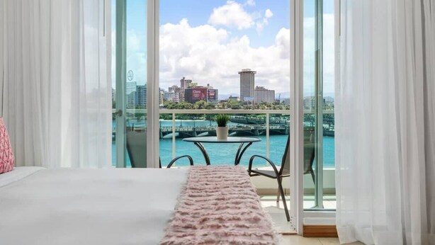 A bedroom with a large bed, a view of a cityscape through a glass door, and a balcony with a table and chairs.