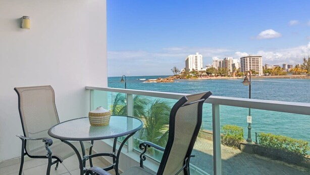 Ein Balkon mit Glastisch und zwei Stühlen, mit Blick auf einen Strand, Gebäude und einen klaren Himmel.