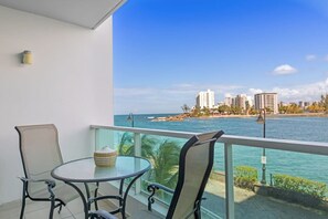 A balcony with a glass table and two chairs, overlooking a beach with buildings and a clear sky.