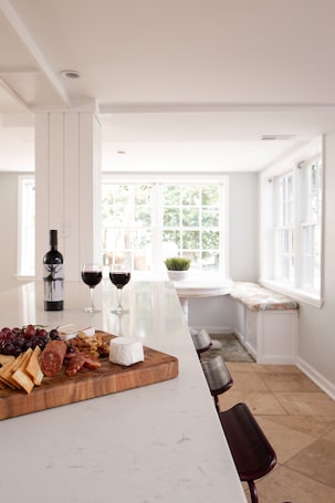 A kitchen with a marble countertop, a wine bottle, two glasses of red wine, a cheese board with various cheeses and grapes, and a window with a view of greenery.