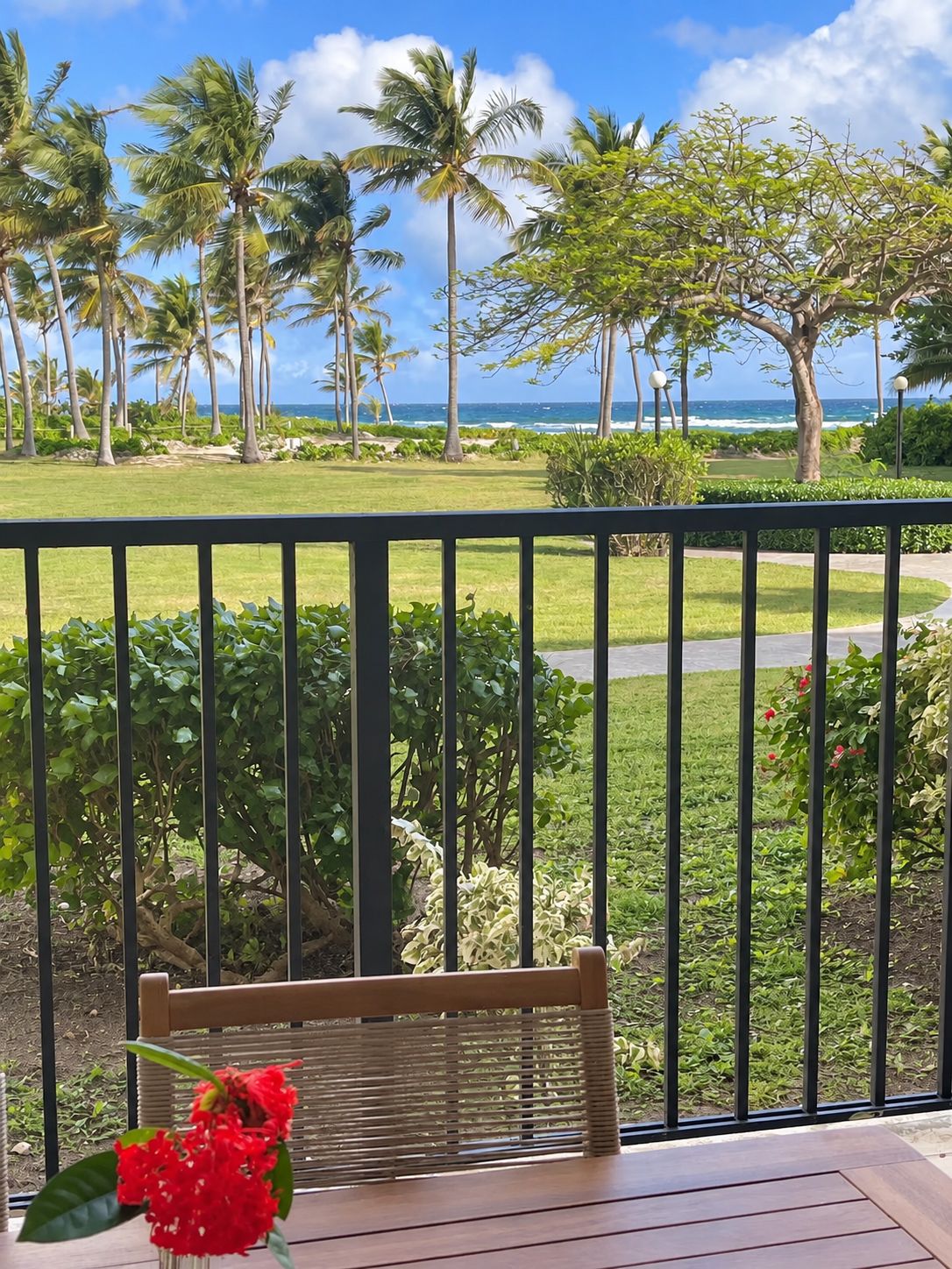 Un balcon avec vue sur l’océan, des palmiers et une chaise en bois ornée d’une fleur rouge.