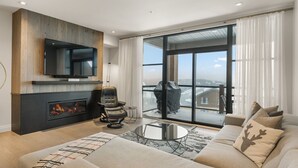 A modern living room with a fireplace, a sofa, a TV mounted on a wooden wall, and a large sliding glass door leading to a balcony. - Tremblant Prestige - Horizon 2-204 (Mont-Tremblant)