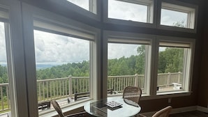 A room with a glass table surrounded by wicker chairs, large windows with a view of a forested landscape, and a wooden deck. - Creekview Manor (Burkesville)