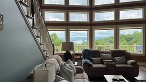 A spacious living room with a large window, a staircase, and a view of greenery. - Creekview Manor (Burkesville)