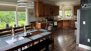 A spacious kitchen with wooden cabinets, a dining table with chairs, and a large window providing a view of greenery. - Creekview Manor (Burkesville)