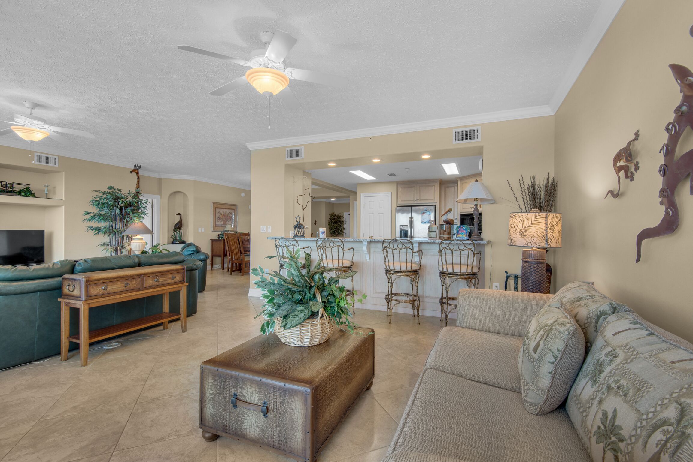 A spacious living room with a green sofa, a beige sofa, a wooden coffee table, and a vintage suitcase. There is a kitchen area with bar stools and a refrigerator.