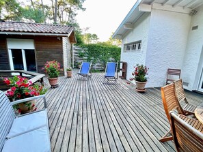 Terraza de madera con sillones y una mesa, rodeada de plantas en macetas y un edificio con fachada blanca.