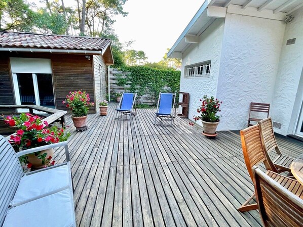 Eine Holzterrasse mit Liegestühlen und einem Tisch, umgeben von Kübelpflanzen und einem Gebäude mit weißer Fassade.