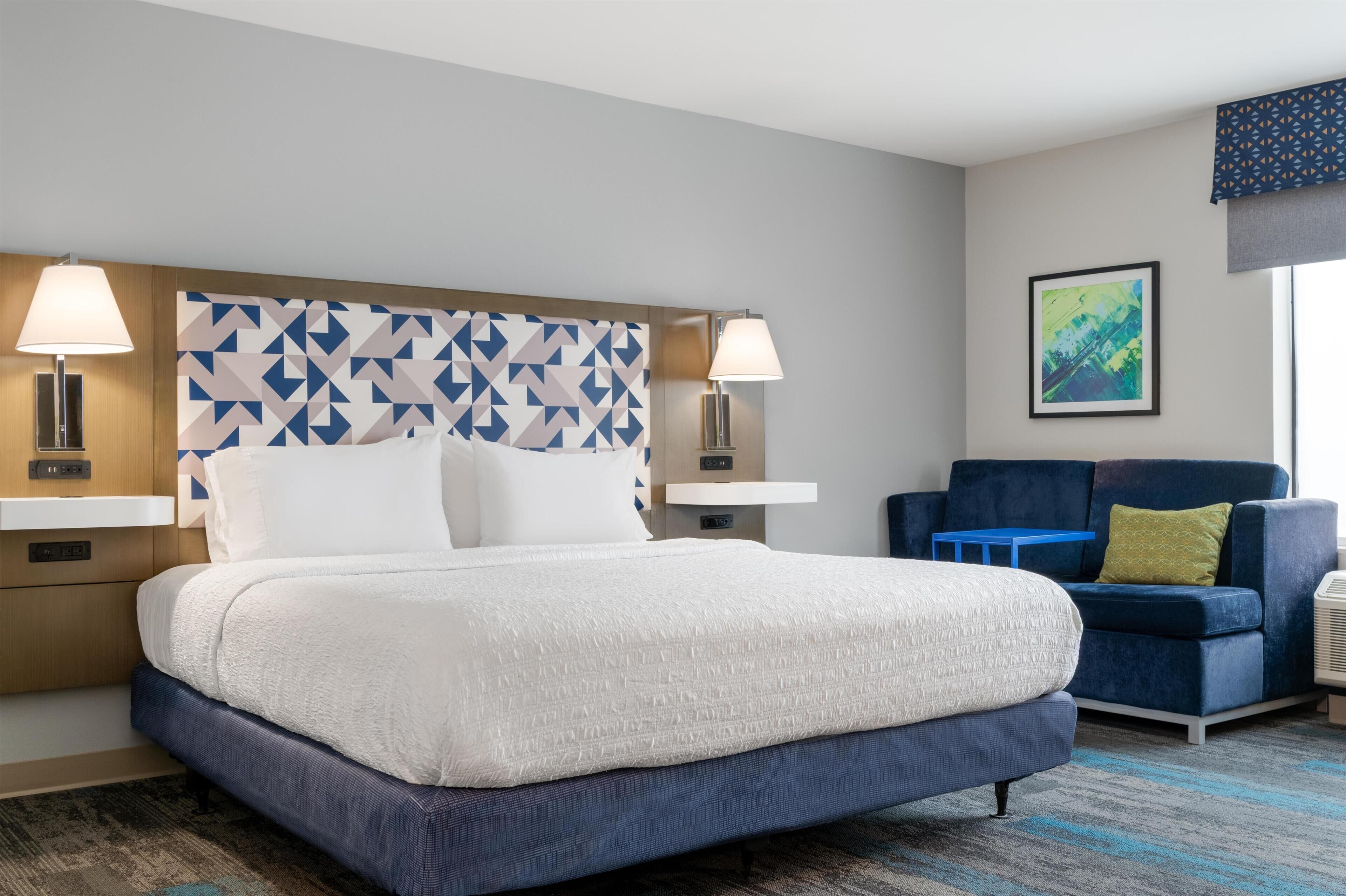 A hotel room with a large bed, two bedside lamps, a blue sofa, a framed artwork, and a window with a view.