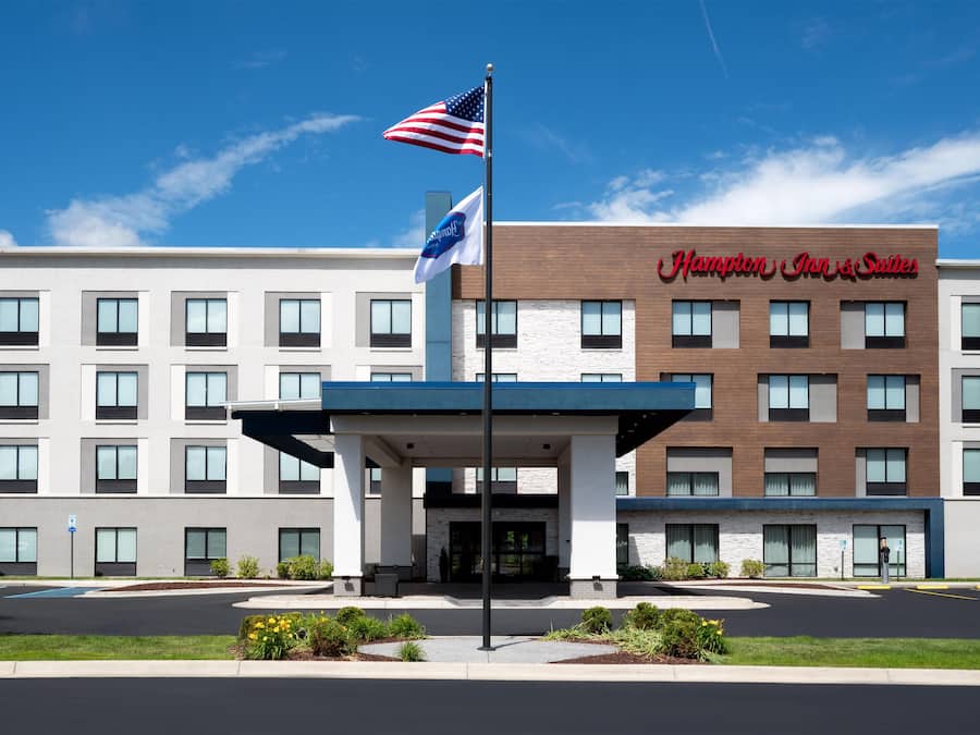 A Hampton Inn & Suites hotel with a modern facade, a flagpole with the American flag, and a clear blue sky.