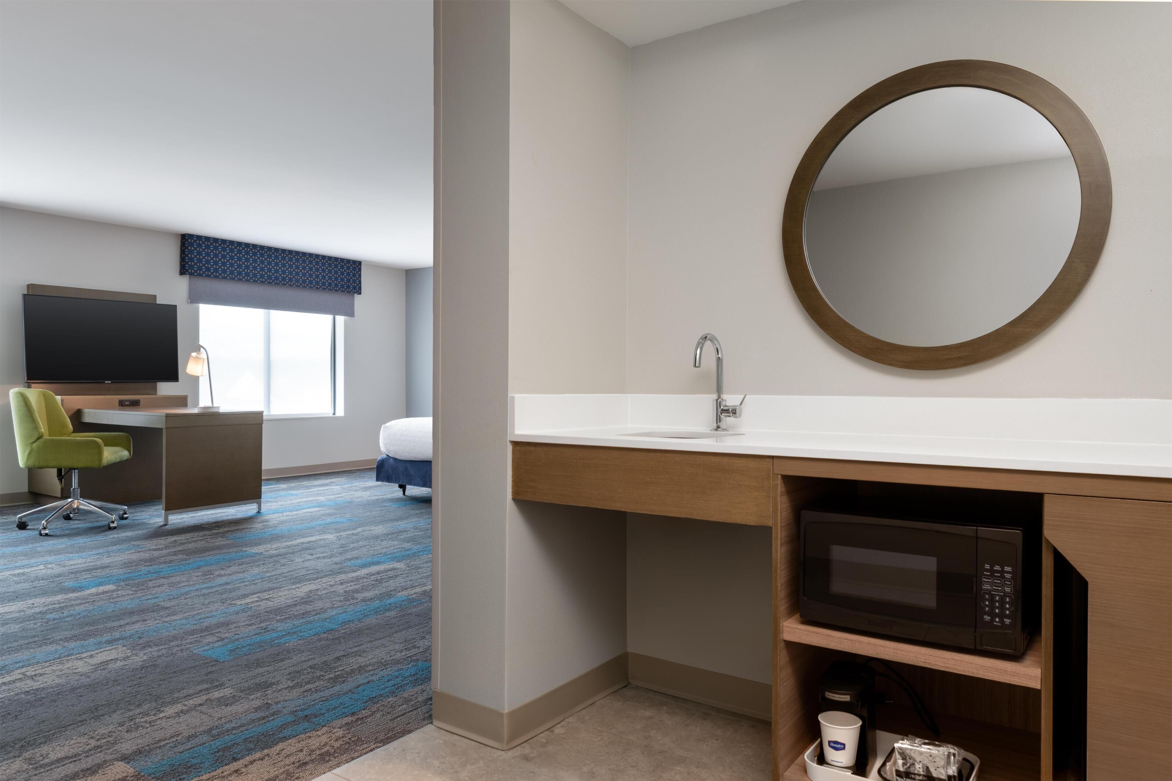 A hotel room with a flat-screen TV, a desk with a chair, a bed, a bathroom with a sink and mirror, and a microwave.