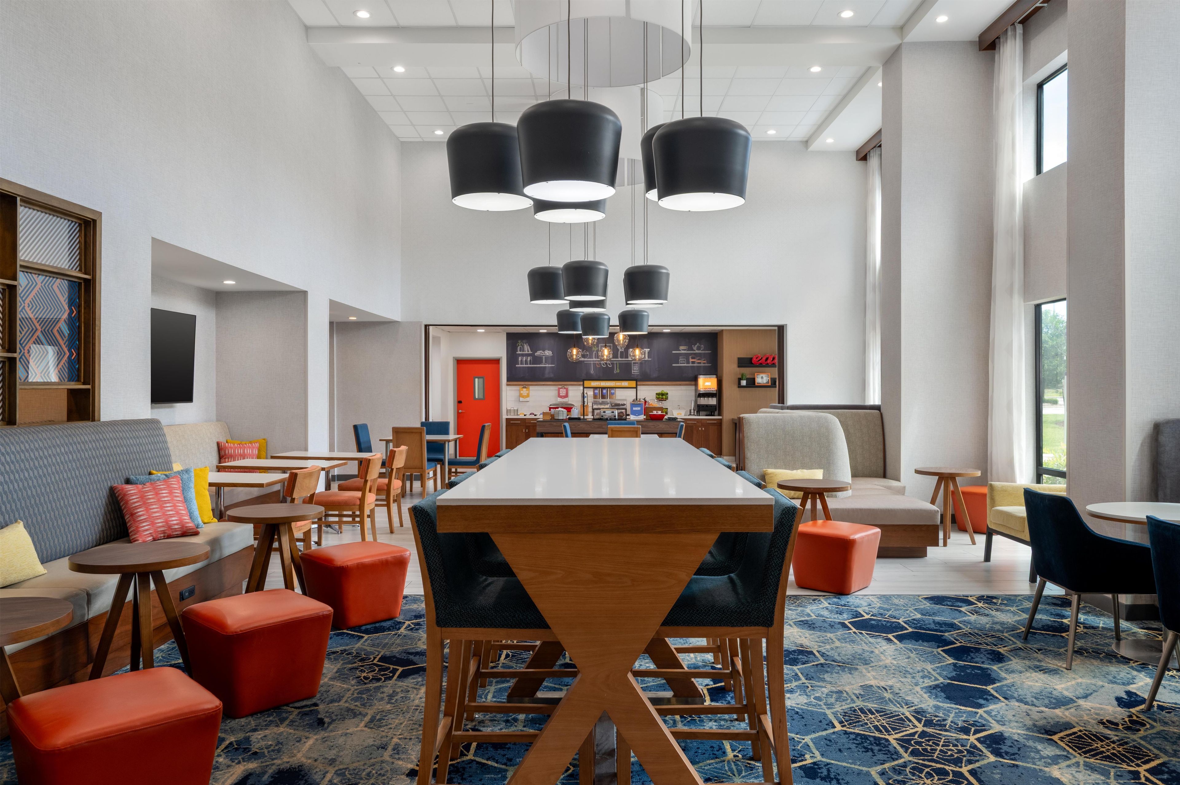 A modern hotel lobby with a long dining table, multiple seating options, and a kitchen area in the background.