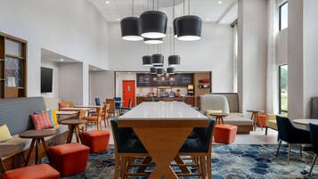 A modern hotel lobby with a long dining table, multiple seating options, and a kitchen area in the background.