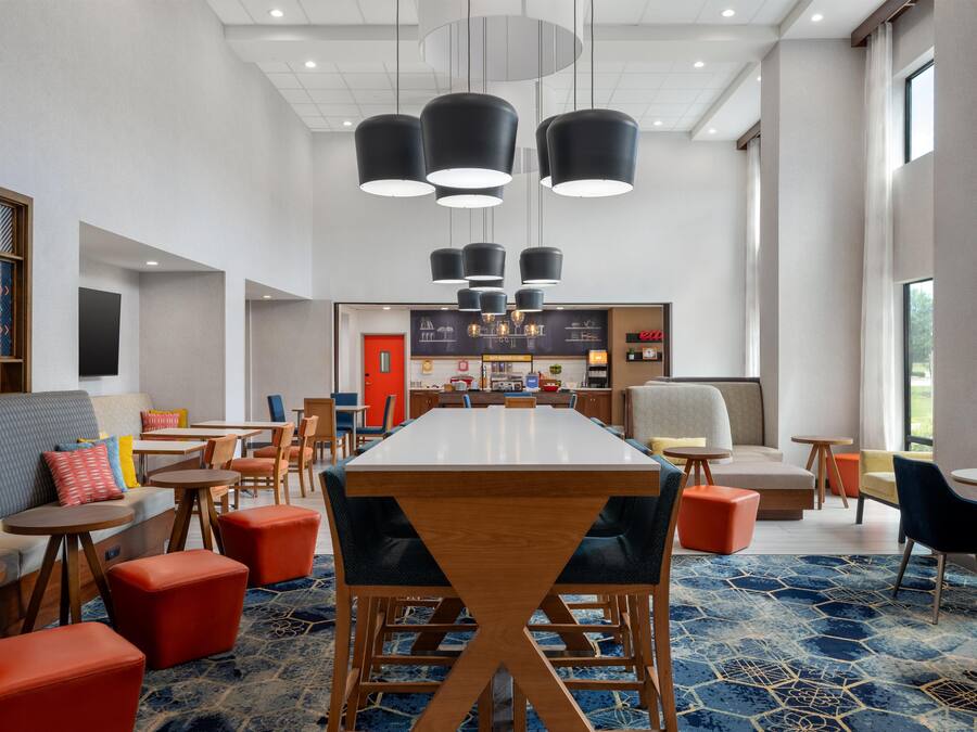 A modern hotel lobby with a long dining table, multiple seating options, and a kitchen area in the background.