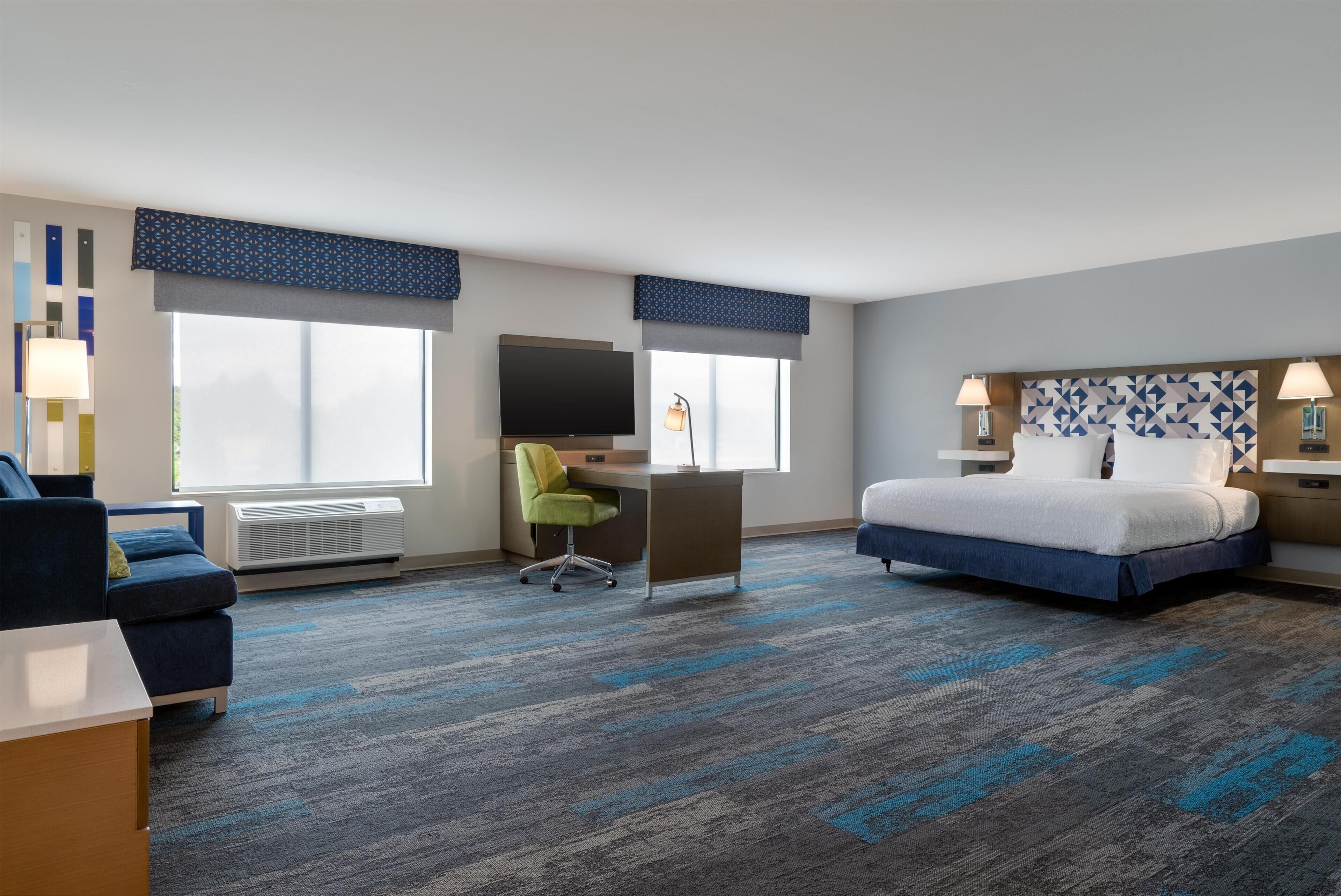 Ein Hotelzimmer mit Bett, Schreibtisch, Stuhl, Fernseher und zwei Fenstern mit Jalousien.