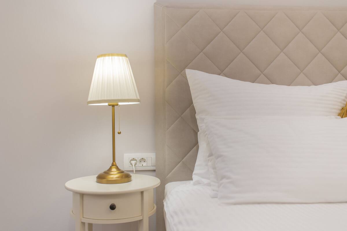 A neatly made bed with a tufted headboard, a bedside table with a lamp, and a power outlet on the wall.