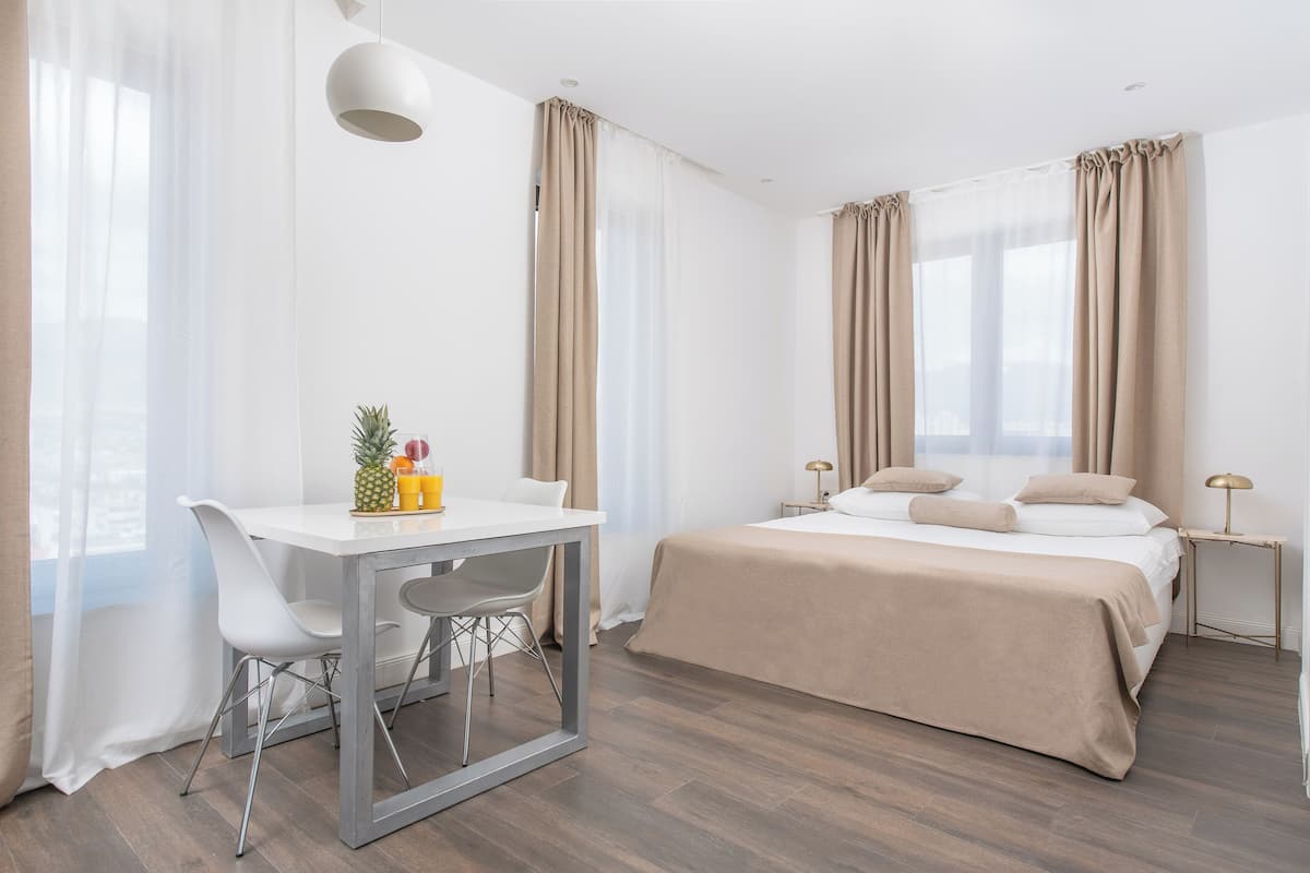 A hotel room with a bed, a desk, and a chair. There is a window with curtains, a lamp, and a small table with fruits.
