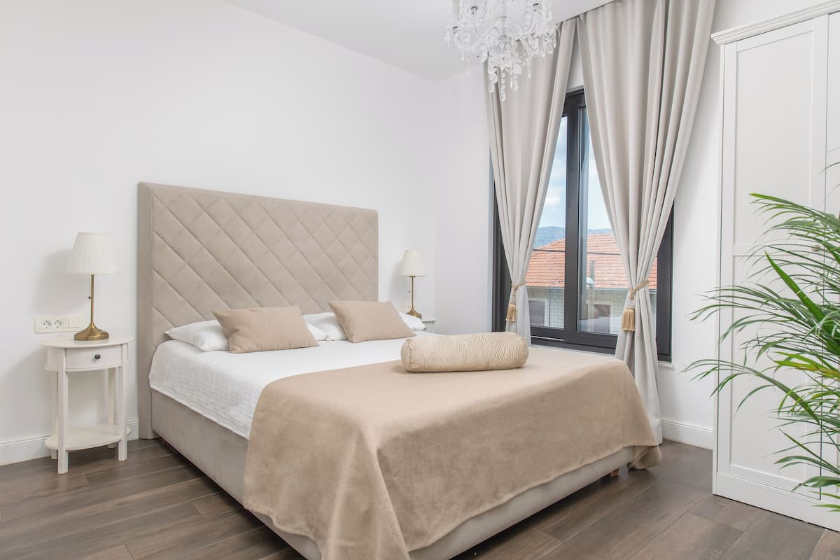 A bedroom with a large bed, a nightstand, a lamp, a chandelier, and a window with curtains.